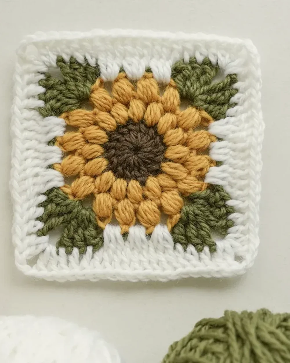 A puff-stitch sunflower granny square in gold, avocado and cream, lying on the porch glider in afternoon light